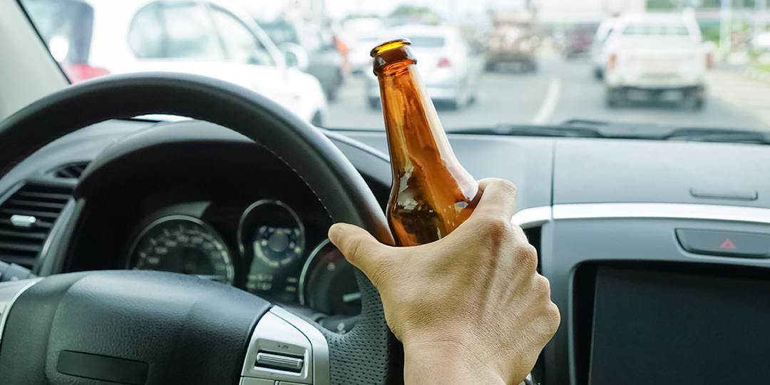 A driver holding a beer bottle in their hand against the steering wheel while in traffic.