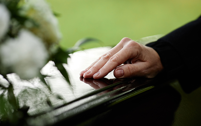 A widow's hand resting on the surface of a dark casket at a funeral with flowers nearby.