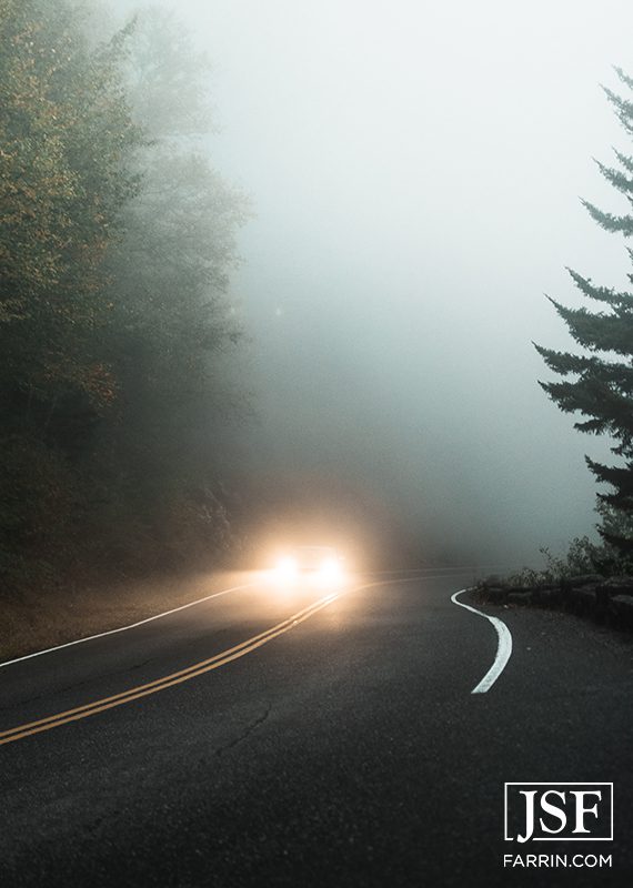Car on a road in a misty forest with its headlights on.