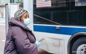 A woman wearing a mask and gloves and a winter coat waiting for a bus at a bus stop.