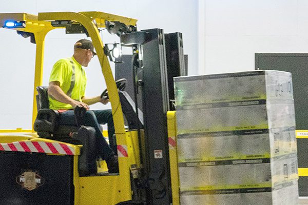 Man in a high vis shirt driving a yellow forklift carrying a pallet in a warehouse.