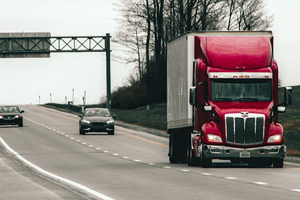 A red tractor trailer on the road in front of two black cars.