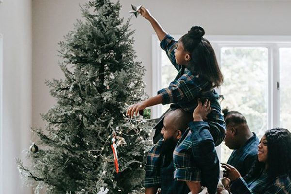 Black man holding his daughter on his shoulders to put a star on the Christmas tree.