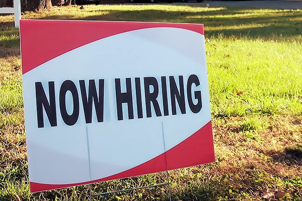 Red and white "Now Hiring" sign in the grass.
