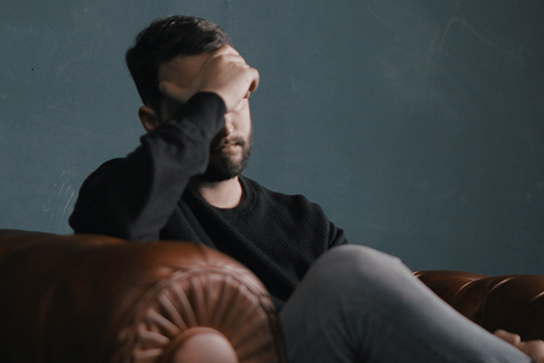A depressed man holding his head in his hand, sitting on a couch in a dim room.