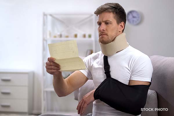 Man using neck brace and arm sling sits on a couch frowning at a Workers’ Compensation denial letter