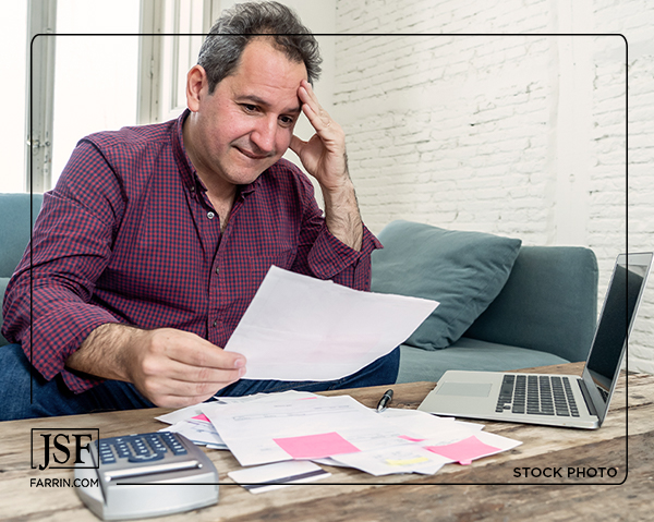 A man sits looking exasperated over a laptop, a pile of bills, and a calculator