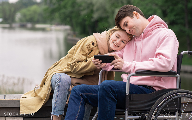 A man in a wheelchair showing his phone to a woman on a bridge outdoors.