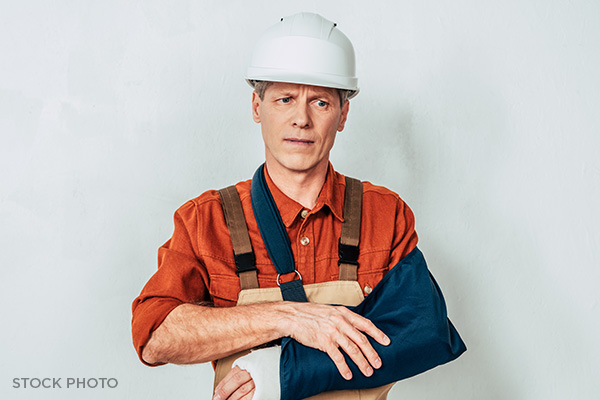 A worker wearing a hard hat and holding his arm in a sling with a worried expression.