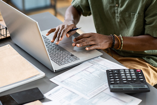 A Black man working on a laptop, typing on the keyboard with forms & a calculator.