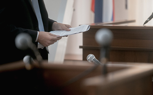 A man reading from documents in a courtroom, with microphones blurred in the foreground.
