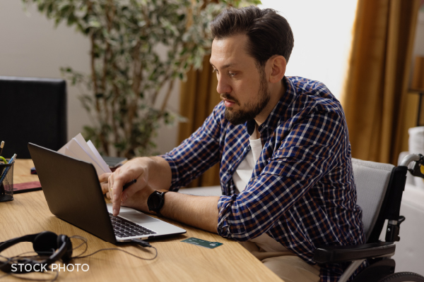 A disabled young man in a wheelchair doing financial calculations on a laptop.