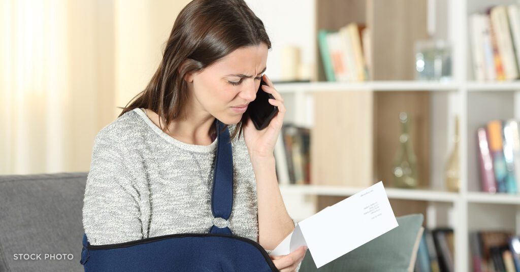 Law Offices of James Scott Farrin workers’ compensation attorneys. An injured woman with her arm in a sling looks concerned while reviewing paperwork on the phone, suggesting a denied or disputed claim.