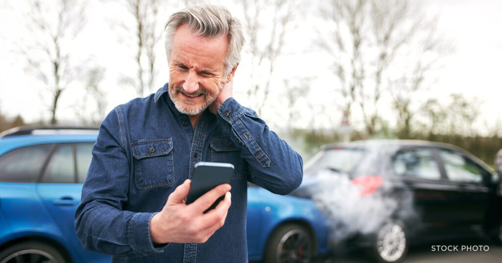 A man making a phone call while rubbing his sore neck wincing in pain after a car crash. James Scott Farrin, auto accident attorneys.